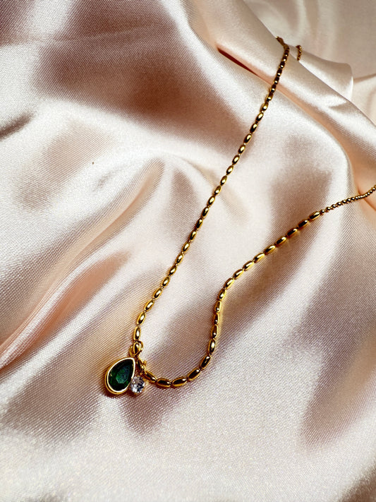 Gold necklace with a green teardrop pendant on a soft pink fabric background